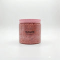 Donatella glow-boosting body scrub in a clear jar with a pink ribbed lid. The scrub is pink with visible exfoliating particles.